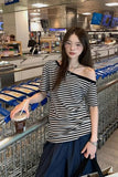 A woman wearing a black and white striped off-the-shoulder t-shirt and dark blue pants stands in a supermarket holding a drink, with shopping carts in the foreground.