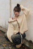 Person wearing a beige cardigan over a black top, sitting against a white wall.