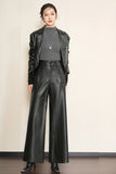 A woman modeling a black faux leather jacket, matching wide-leg pants, a dark gray mock neck top, and black pointed-toe boots against a plain beige wall.