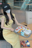 Woman in a yellow dress and black cap sitting at a table with a tray of food in an indoor setting.