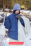 Person wearing a blue coat, beanie, and gloves in a snowy forest setting with a visible brand logo.
