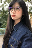 A close-up portrait of a young woman with long black hair wearing round metal-framed eyeglasses with purple-tinted lenses and a dark blue denim jacket, photographed outdoors with trees and a railing in the background.