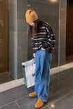 Person wearing a striped sweater, blue jeans, and yellow beanie standing against a tiled wall.
