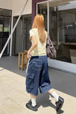 Person wearing a yellow top and blue cargo shorts walking outside a building.