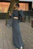 Woman in a long dark gray dress standing on a wooden deck with a tree and building in the background.