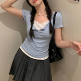 Person wearing a light blue top with lace details and a black skirt, standing against a plain wall.