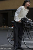 Woman standing next to a bicycle with a black basket, wearing a white sweater and dark pants.