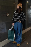 Person wearing a striped sweater, beanie, and jeans holding a green bag against a dark tiled wall.