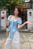 Woman in a light blue off-shoulder top and white pants holding a drink outdoors.