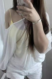 A person taking a mirror selfie wearing a white sheer long-sleeve top over a pale yellow camisole, paired with white shorts and a silver bracelet.