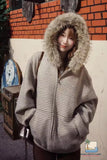 Person wearing a gray hooded coat with fur trim in front of a brick wall.