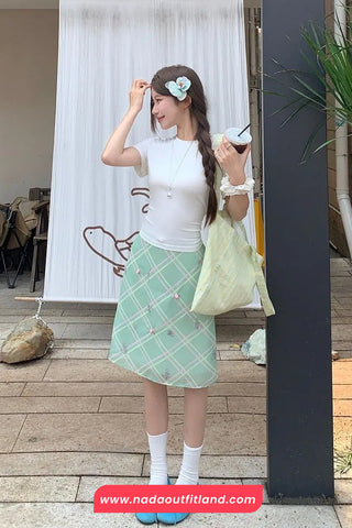 Woman in a white top and green checkered skirt standing outdoors with a plant and banner in the background.