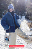 Person in winter clothing with a blue coat and white bag in a snowy forest, brand 'www.nadaoutfitland.com' visible.