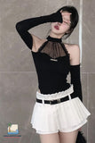 Person wearing a black long-sleeve top with sheer details and a white pleated skirt against a tiled wall.