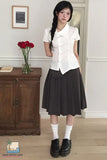 Woman in a white shirt and black skirt standing in a room with a wooden cabinet and flowers.