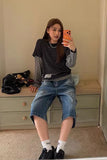 A woman takes a mirror selfie wearing a black and white striped t-shirt layered over a gray long-sleeve knit top, paired with blue denim shorts, white socks, and black sneakers. She is sitting on a light-colored wooden dresser in a room with beige walls.