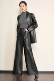 A woman modeling a black faux leather outfit consisting of a cropped blazer, wide-leg trousers, a dark gray mock neck top, and black high-heeled shoes, standing against a light beige wall.