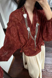 Person wearing a red patterned blouse with long sleeves and a necklace indoors.