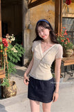 A woman wearing a beige short-sleeve V-neck knit top and a dark denim mini skirt, standing outdoors in front of a rustic building with potted plants and wooden beams.