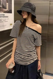 A woman wearing a black and white horizontally striped off-the-shoulder t-shirt, a dark gray bucket hat, and a dark skirt, holding a coffee cup in an elevator setting.