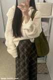 Person wearing a cream sweater and plaid pants with a green bag, taking a mirror selfie.