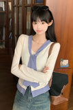Woman wearing a layered outfit with a sweater over a checkered shirt, standing indoors.