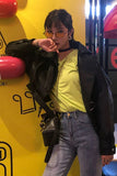 Person wearing a black leather jacket, yellow shirt, and blue jeans against a bright yellow background.