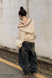 Person wearing a beige oversized sweater and dark jeans standing against a light-colored wall.