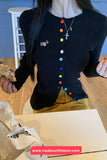 Person wearing a black cardigan with colorful buttons, sitting on a wooden floor.