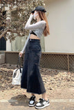 A woman wearing a gray long-sleeve crop top and a dark blue denim maxi skirt, standing outdoors on a paved area with dry leaves. She is wearing a black cap, a white face mask, and black-and-white sneakers, and holding a white handbag.