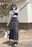 A woman wearing a gray long-sleeve crop top, a black crop top underneath, a dark gray denim maxi skirt, a black baseball cap, and a white face mask, holding a white tote bag and posing outdoors.