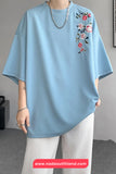 Light blue t-shirt with floral embroidery worn by a person, with a gray background and website watermark.