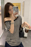 A woman takes a mirror selfie wearing a black and white horizontally striped off-the-shoulder t-shirt, holding a black quilted shoulder bag and a iced coffee in a tiled indoor space.