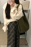 Person wearing a cream sweater and plaid pants with a green bag indoors.