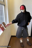 Person wearing a black hoodie, red cap, and gray shorts standing indoors.