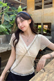 A woman wearing a sheer cream-colored short-sleeve lace-trim top with a crossover front, styled with a blue patterned headband and a silver chain bag strap, set in an outdoor cafe environment.