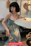 Woman in a floral dress standing in a room with a lamp and table.