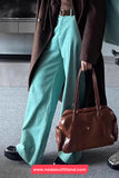 Person wearing a brown coat and teal pants holding a brown leather bag indoors.