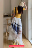 Person wearing a yellow top and white skirt with blue accents, standing in a room.