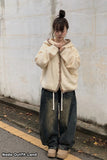 Person wearing a beige cardigan and dark jeans standing against a white wall.
