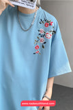 Light blue t-shirt with floral embroidery held by a person against a neutral background.