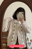 Person wearing a white cardigan with floral patterns in front of a mirror, holding a phone.