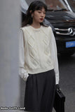 Woman wearing a white cable knit sweater and dark pants, standing on a street.