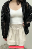 Person wearing a black sheer jacket, white tank top, and beige shorts with a plain background.