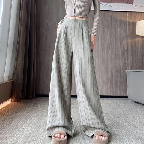 Person wearing light gray ribbed top and wide-leg pants in a room with wooden wall and white curtain.